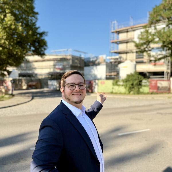 Gilbert Weber in zeigender Pose. Er zeigt auf zwei in Bau befindliche Gebäude, die sich unscharf im Hintergrund befinden.