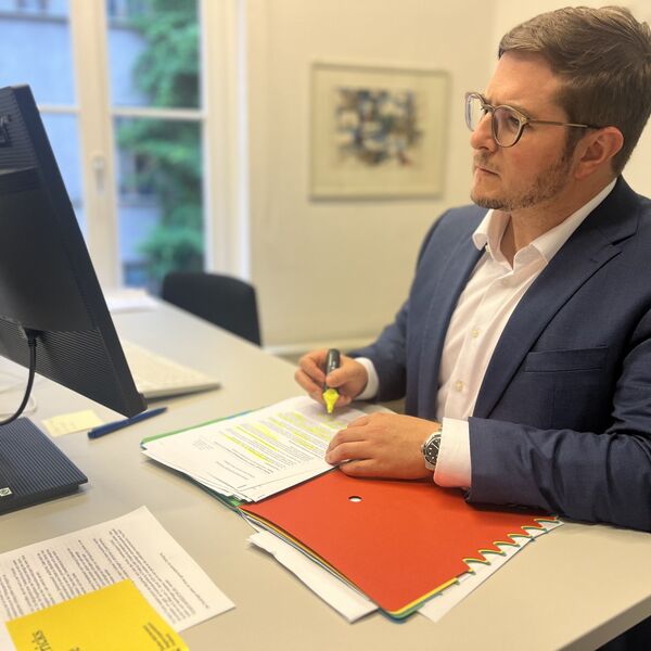 Gilbert Weber sitzt am Arbeitsplatz am Schreibtisch und Computer mit einem gelben Marker ein Dokument in einer Akte markierend.