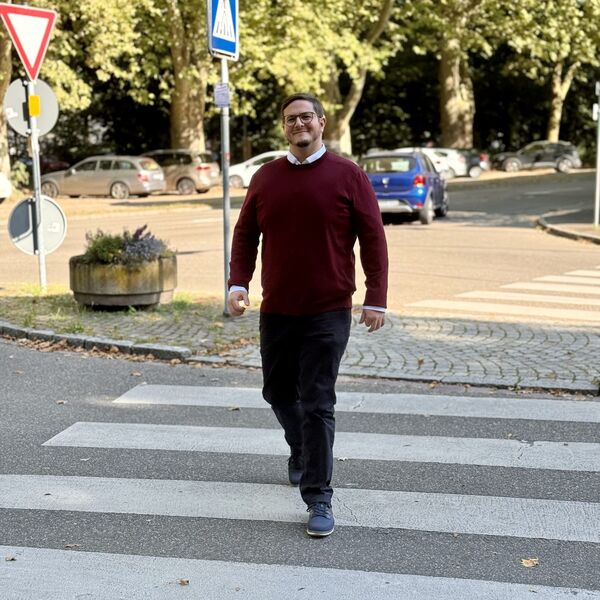 Gilbert Weber geht über einen Zebrastreifen. Im Hintergrund eine Baumallee, ein blaues Auto und zwei Verkehrsschilder