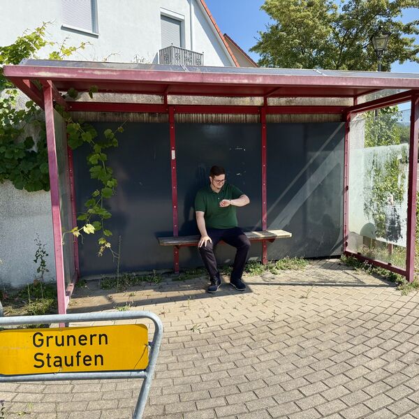 Mann sitzt in einem Bushaltestelle und wartet auf den Bus. Er schaut auf seine Uhr. Im Vordergrund steht ein gelbes Verkehrschild "Grunern, Staufen"